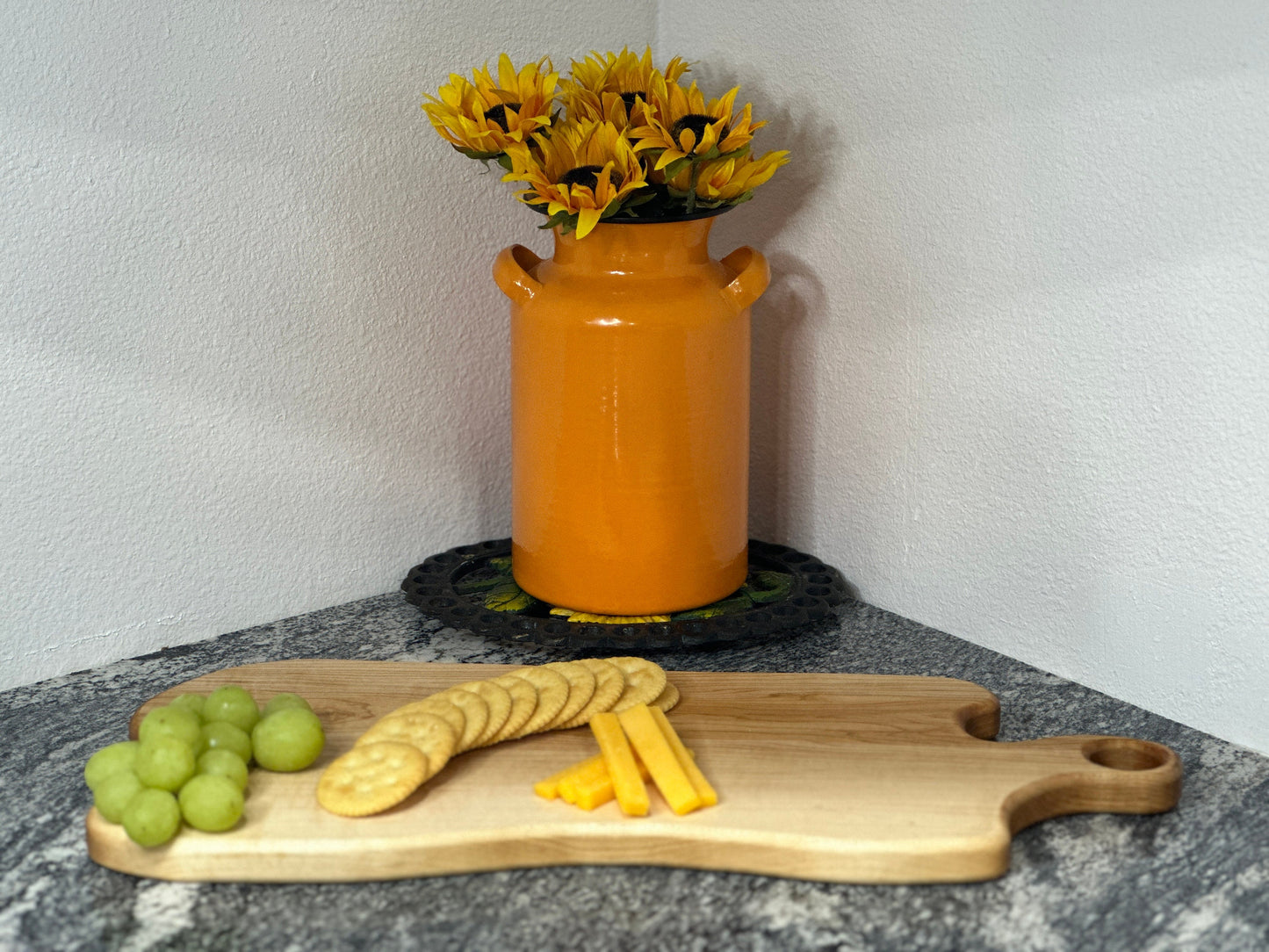 Maple light wood charcuterie board with grapes, cheese and crackers by an orange milk can vase with sunflowers