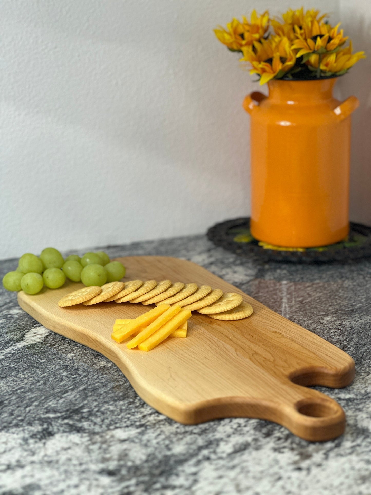 Maple light wood charcuterie board with grapes, cheese and crackers by an orange milk can vase with sunflowers