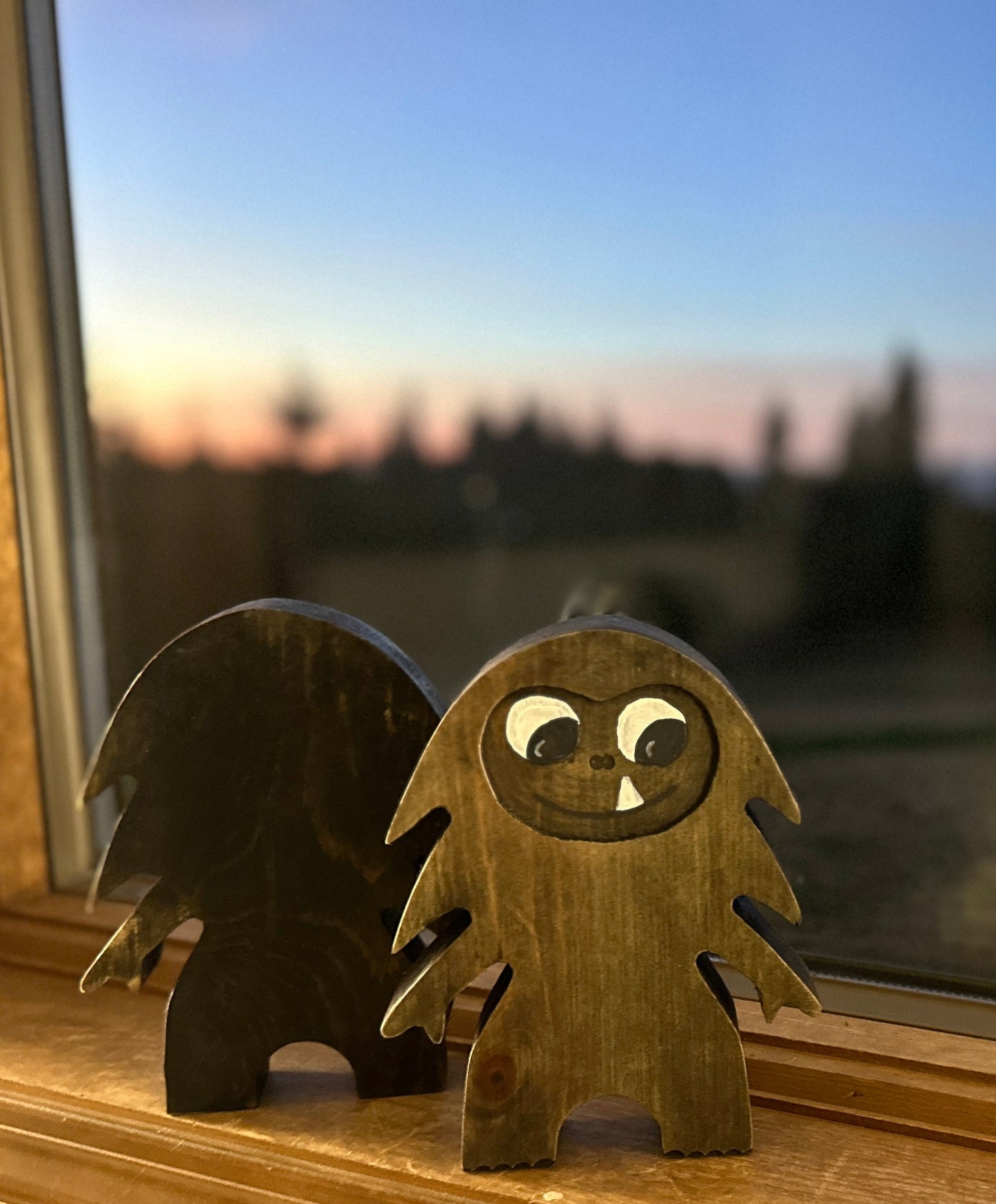 2 Hardwood Bigfoot in blue pine and walnut stain with 1 facing forward smiling one-tooth mouth, and 1 facing backwards showing natural wood grain pattern on windowsill with blurred outdoor view