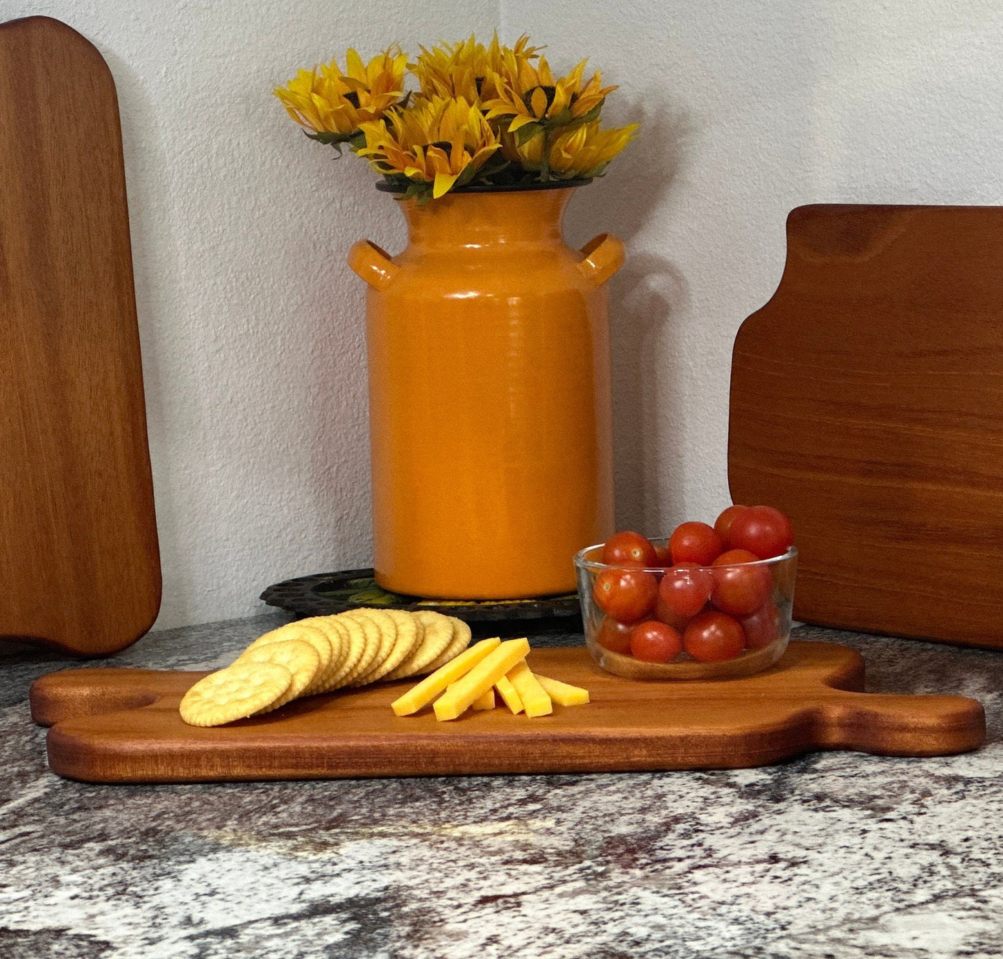 Mahogany red hardwood jigsaw puzzle shaped charcuterie board with cherry tomato, cheese and crackers displayed in front of an orange milk can vase with sunflowers and other cheese boards