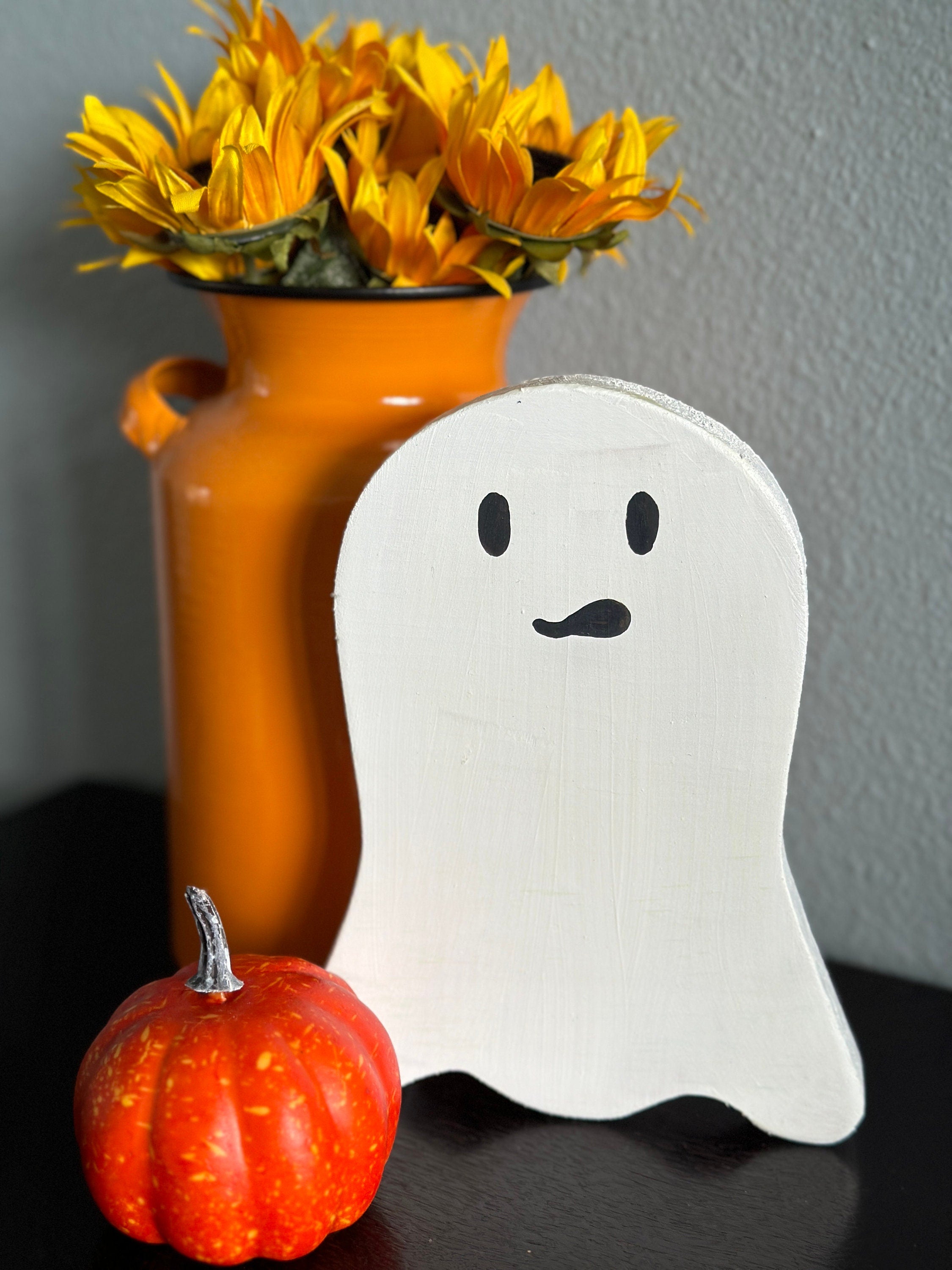 White painted wooden ghost in front of an orange pot of flowers with a small orange pumpkin off to the left side.