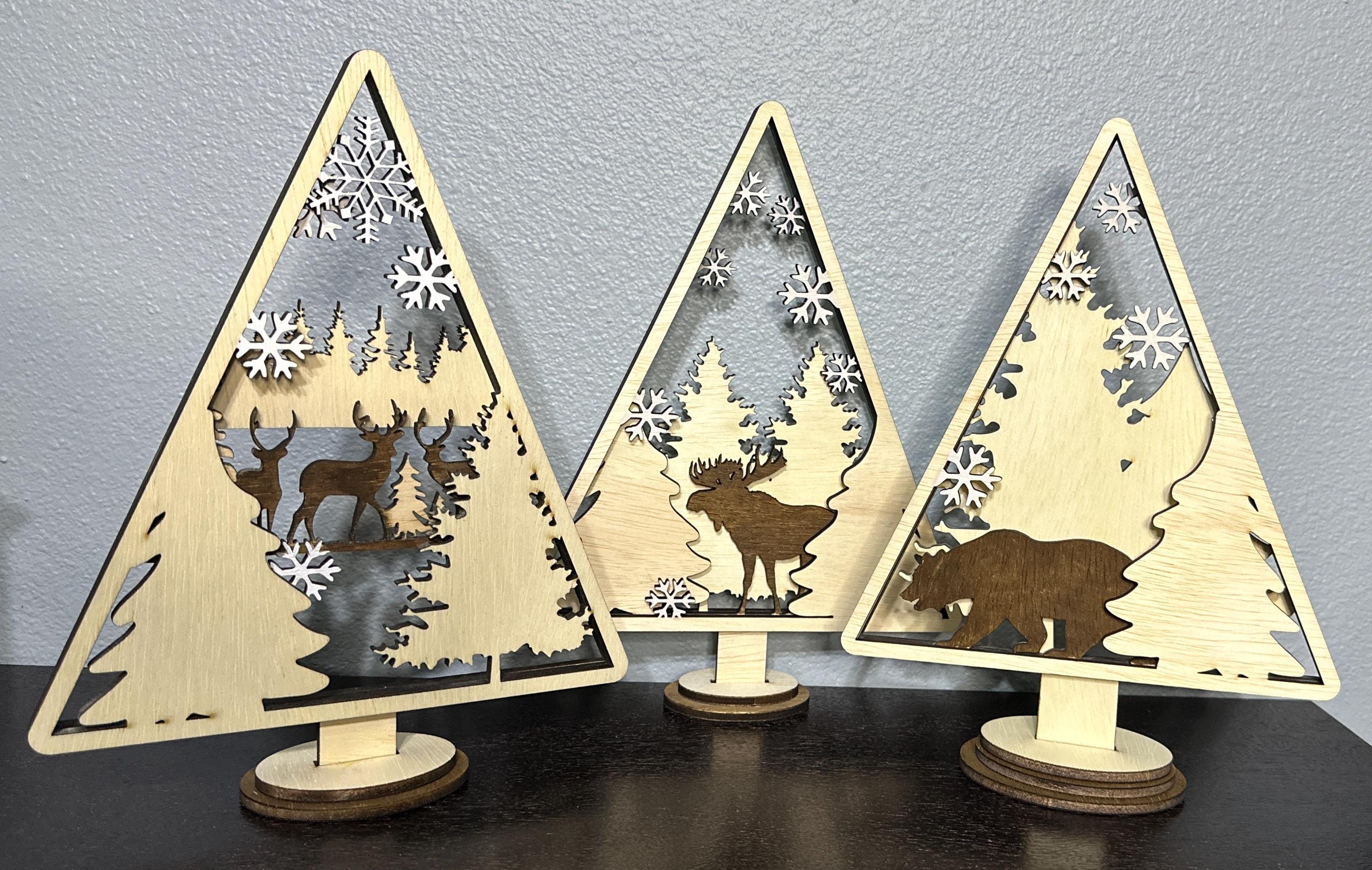 Three wooden layered shelf Christmas decorations.  Each displaying a holiday scene including trees, snowflakes, a  moose, and a bear.