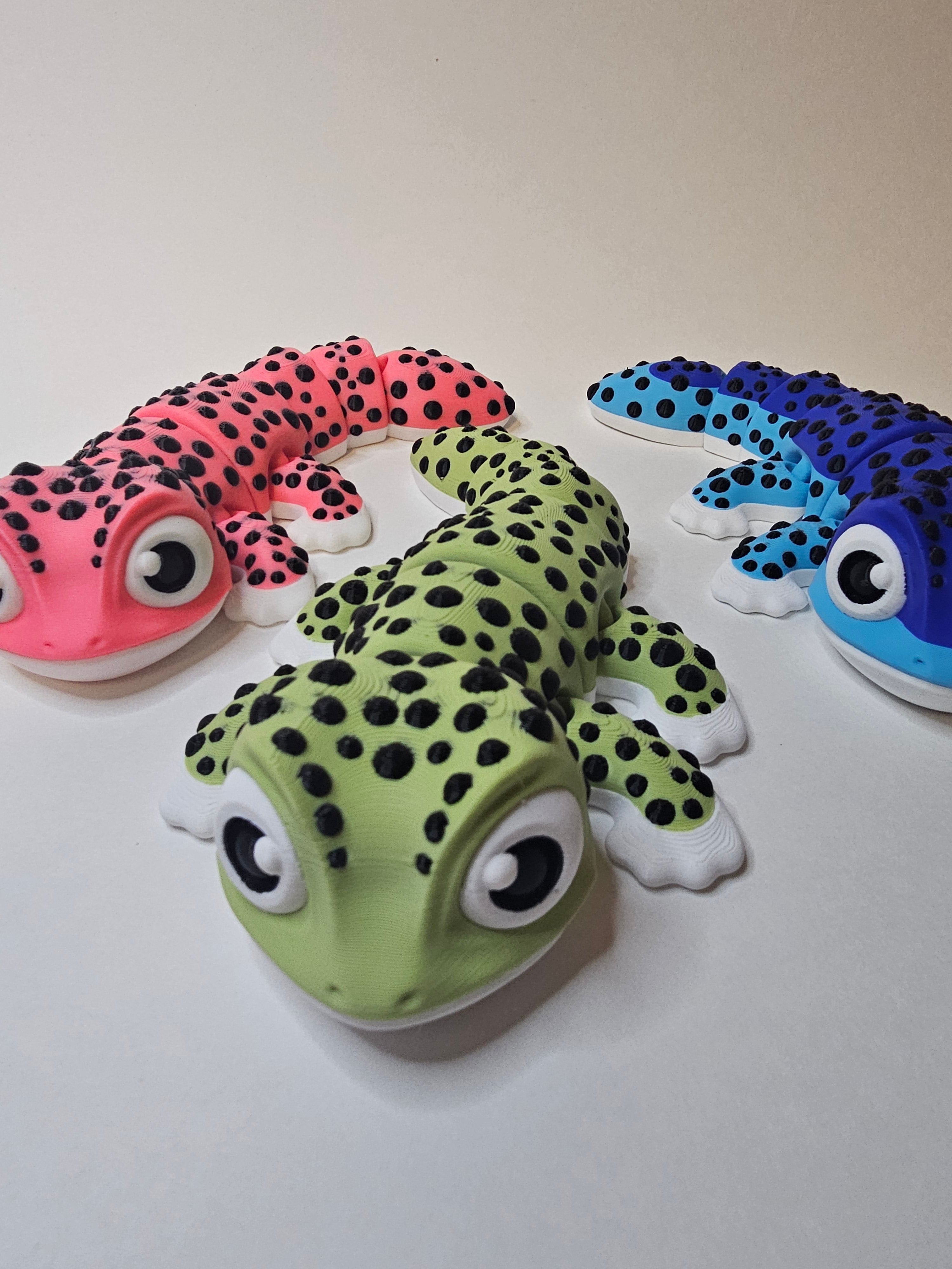 Three cartoon-style gecko toys with large eyes, arranged on a plain light background. One is pink with black spots, one is green with black spots, and one is two-tone blue with black spots.