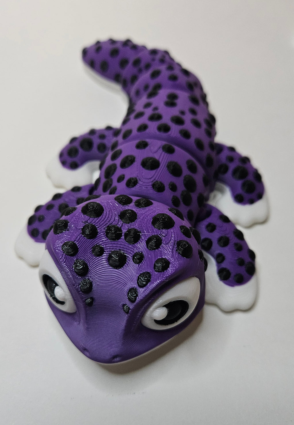 A purple gecko figurine with black spots, white toes, and large cartoon eyes, photographed on a plain white surface.