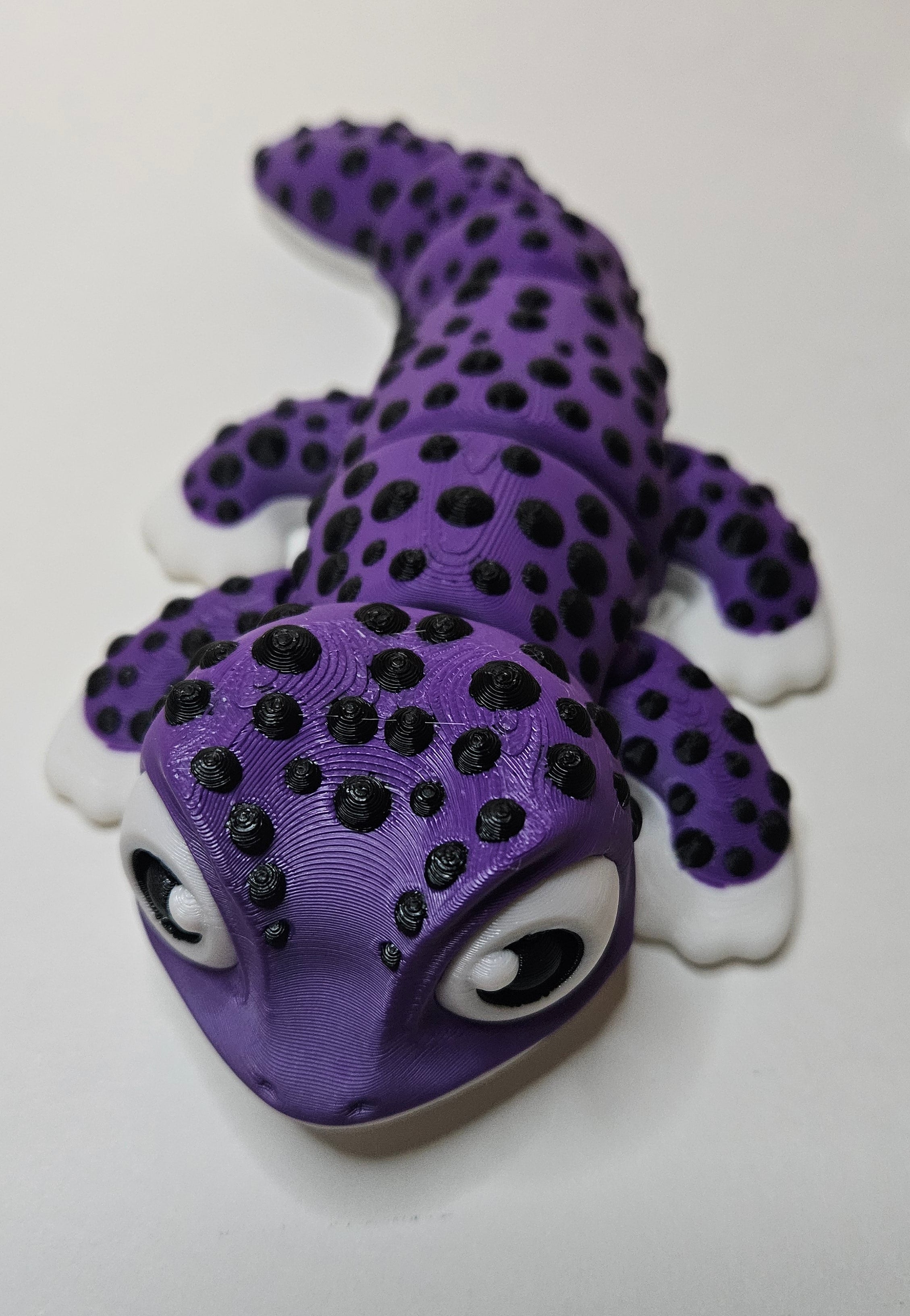 A purple gecko figurine with black spots, white toes, and large cartoon eyes, photographed on a plain white surface.