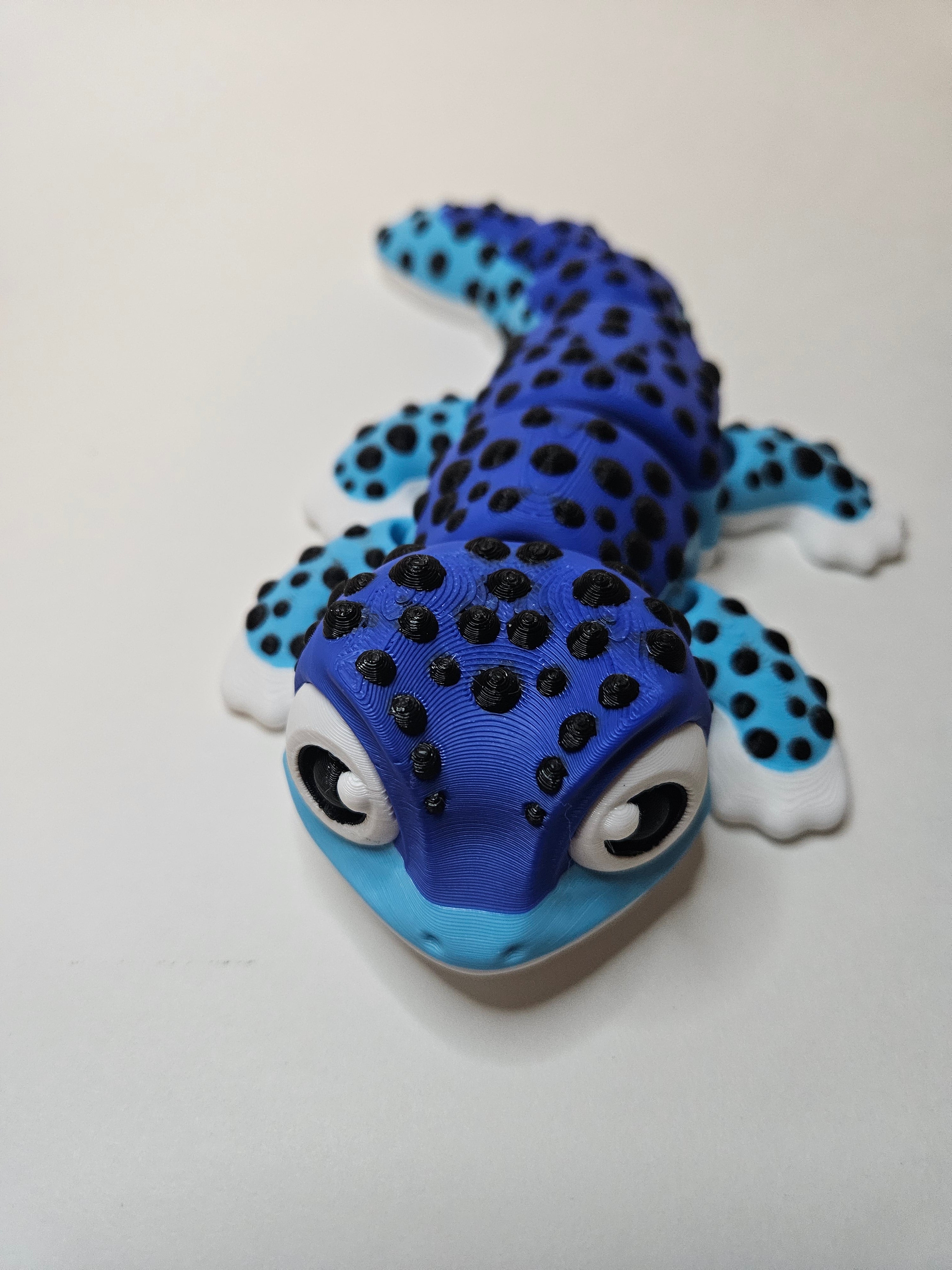 A 3D-printed articulated gecko toy in vibrant two-tone blue, black, and white, photographed on a plain off-white background.
