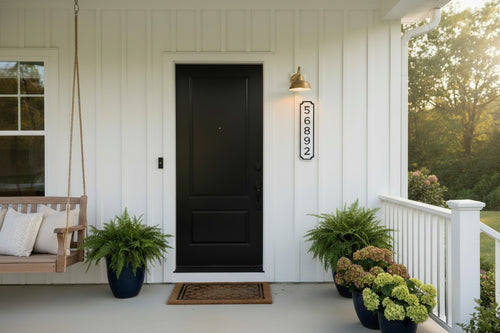 Vertical white and black address sign on a white house with a black door and plants on the porch