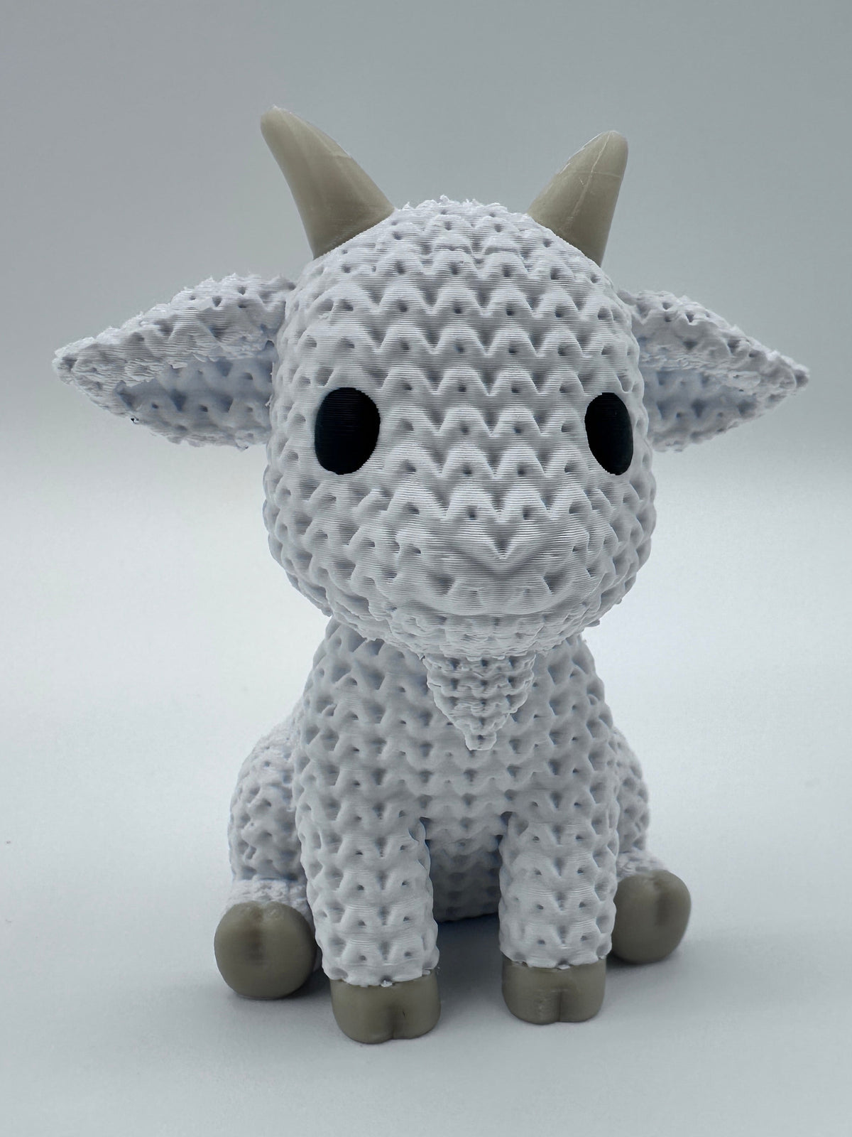 A knit-inspired goat with a white textured body, black eyes, and light brown horns and hooves, photographed against a plain light gray background.