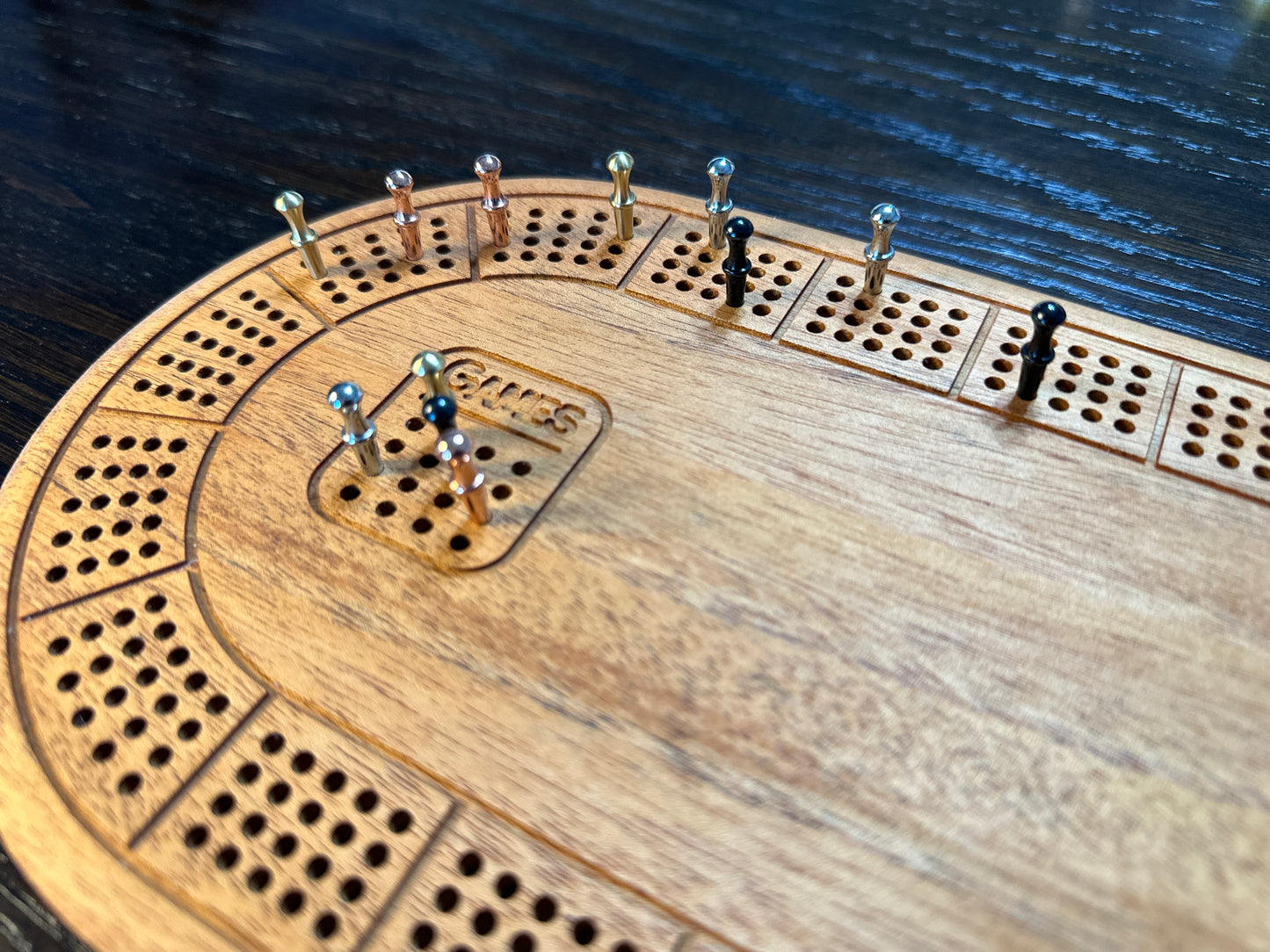 Zoomed in view of oval 4 player hardwood cribbage board with metal pegs in silver, gold, black and rose gold