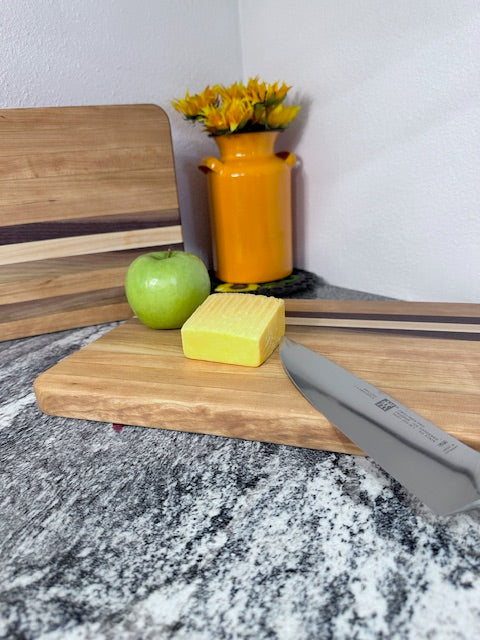 Cherry maple walnut striped hardwood rectangle cutting boards with apple, cheese and knife displayed by an orange milk can vase with sunflowers