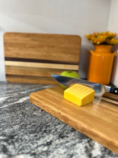 Cherry maple walnut striped hardwood rectangle cutting boards with apple, cheese and knife displayed by an orange milk can vase with sunflowers