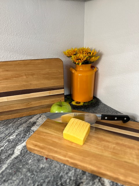 Cherry maple walnut striped hardwood rectangle cutting boards with apple, cheese and knife displayed by an orange milk can vase with sunflowers