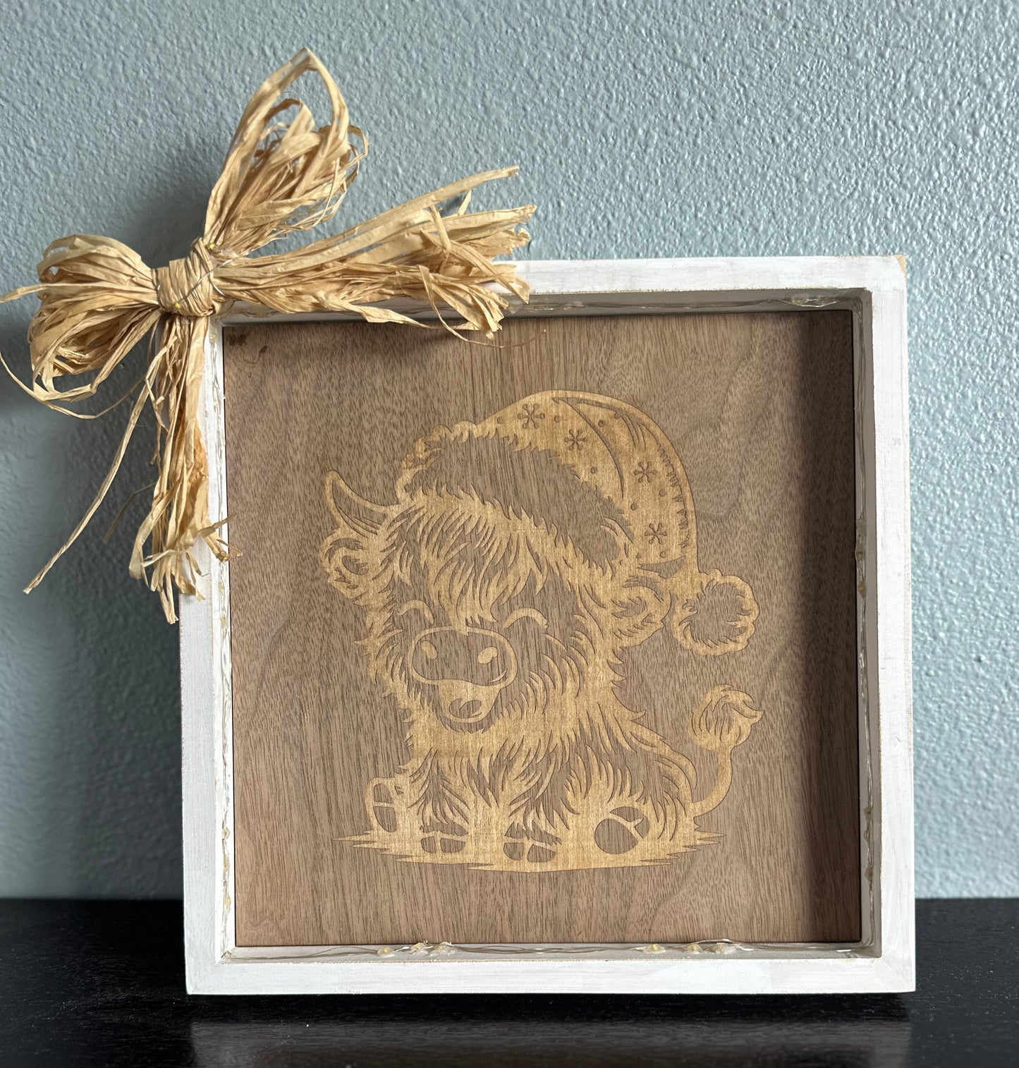Wooden picture frame with engraved design of a highland cow laughing and wearing a Santa hat, accented with a straw corner bow