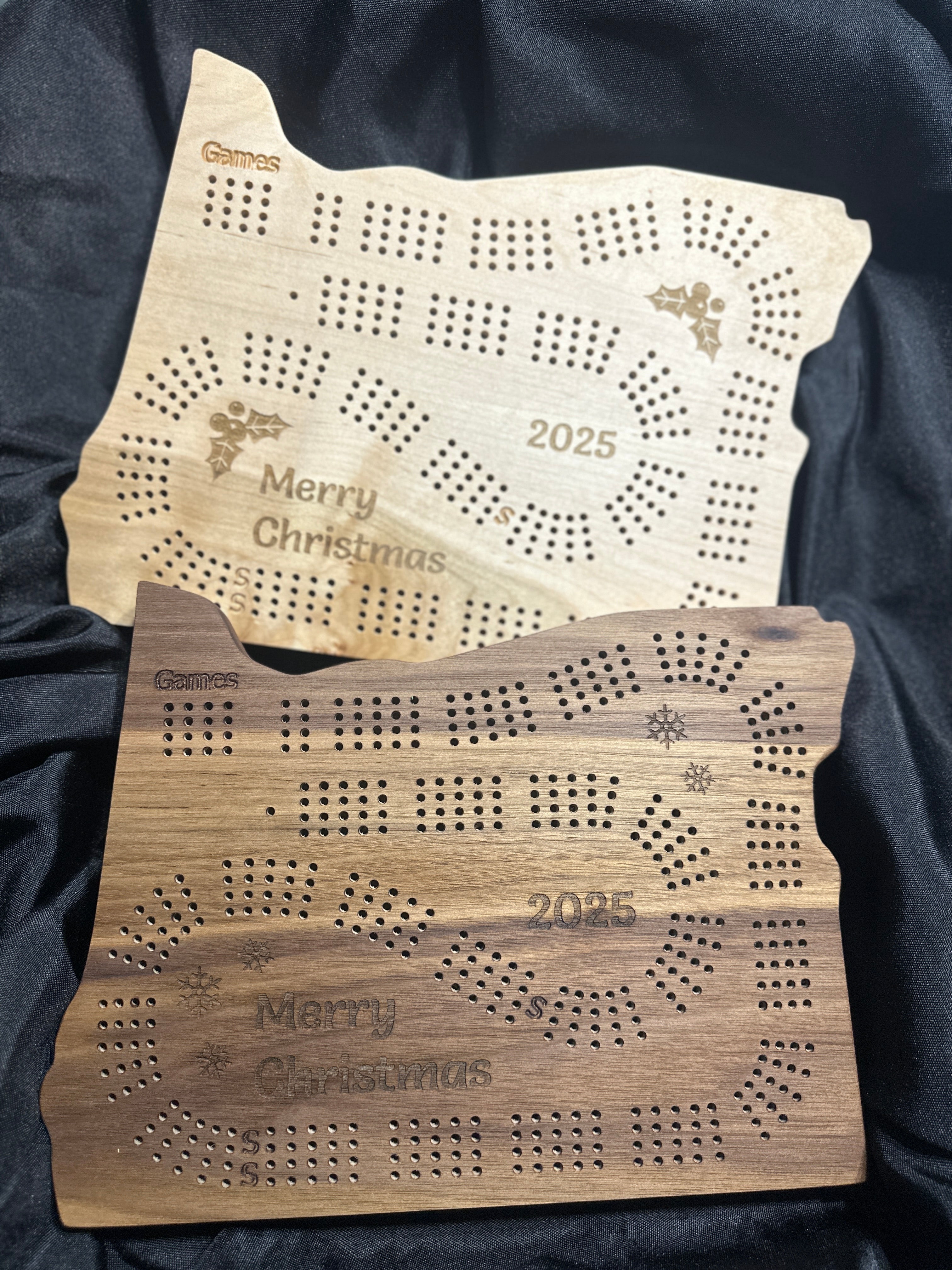 Two wooden Christmas engraved Cribbage boards with 'Merry Christmas 2025' and 'snowflakes and holly' on them on a dark background