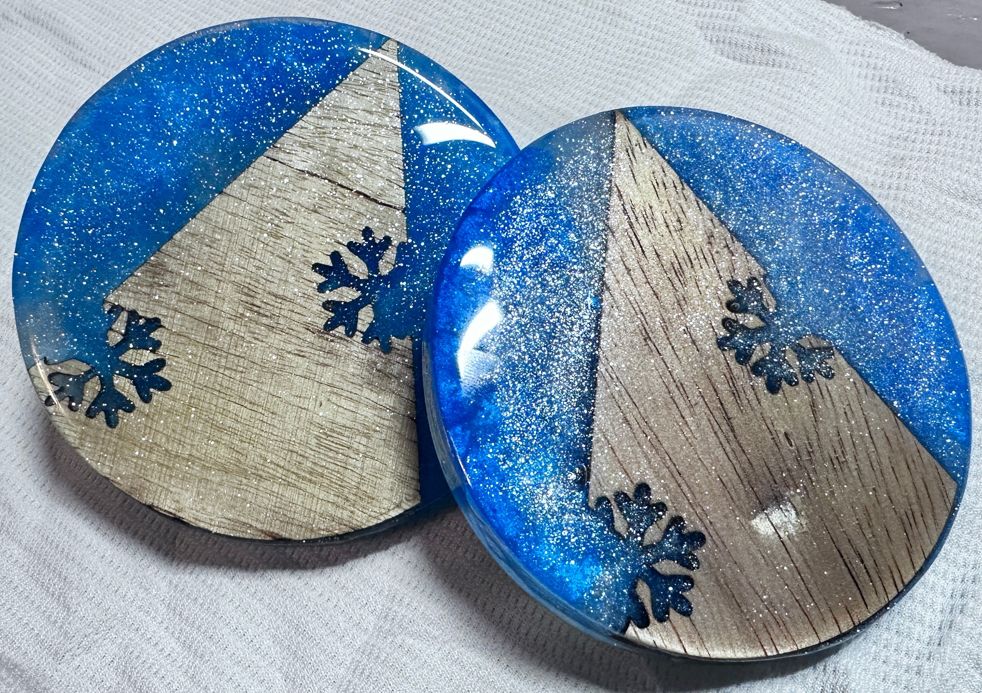 Two round coasters with blue glitter epoxy and wood snowflake designs on a white background.