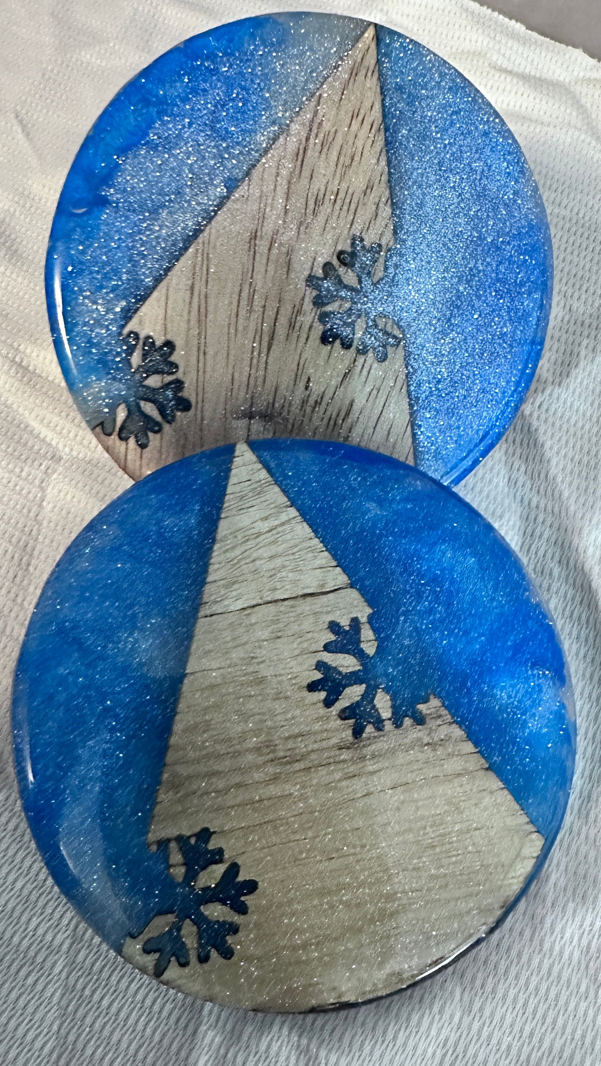 Two round sparkling blue and wood coasters with snowflake designs on a white cloth background.