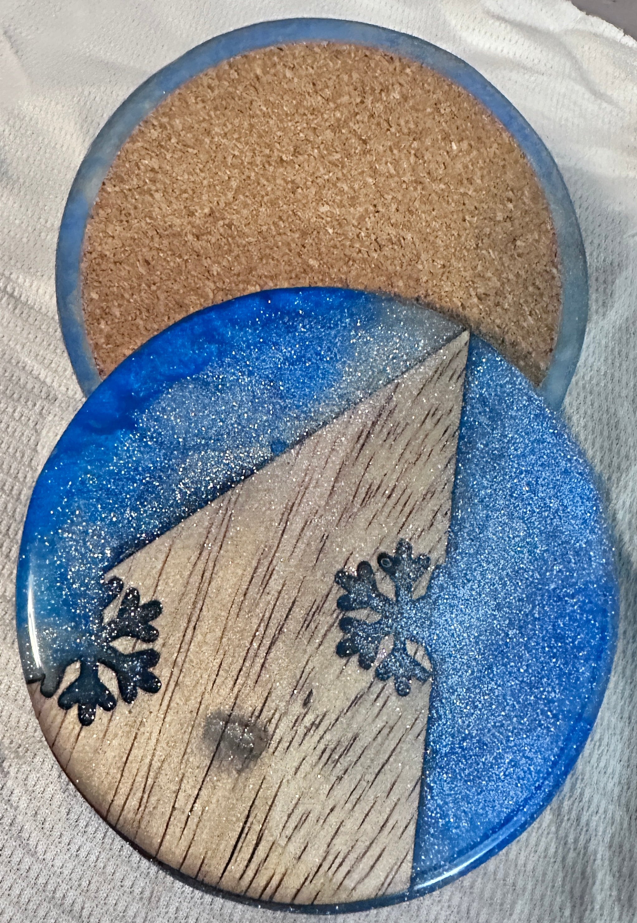Two round coasters with blue rims, cork backings, and sparkling blue snowflake coasters