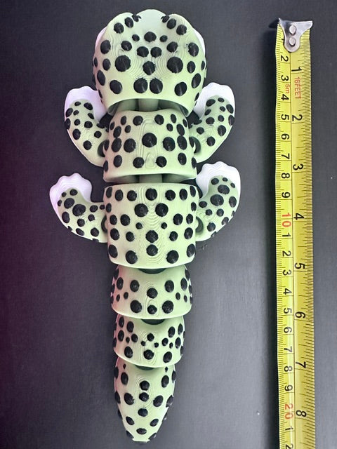 Jumbo green 3D printed articulated Gecko, shown on a dark surface with a yellow measuring tape above it for scale showing just over 8 inches in length. The toy is pale green with black spots and white feet