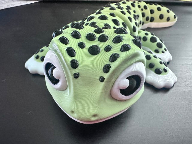 A 3D-printed Jumbo Gecko toy with a light green body, black spots, and large cartoon eyes, resting on a dark surface.