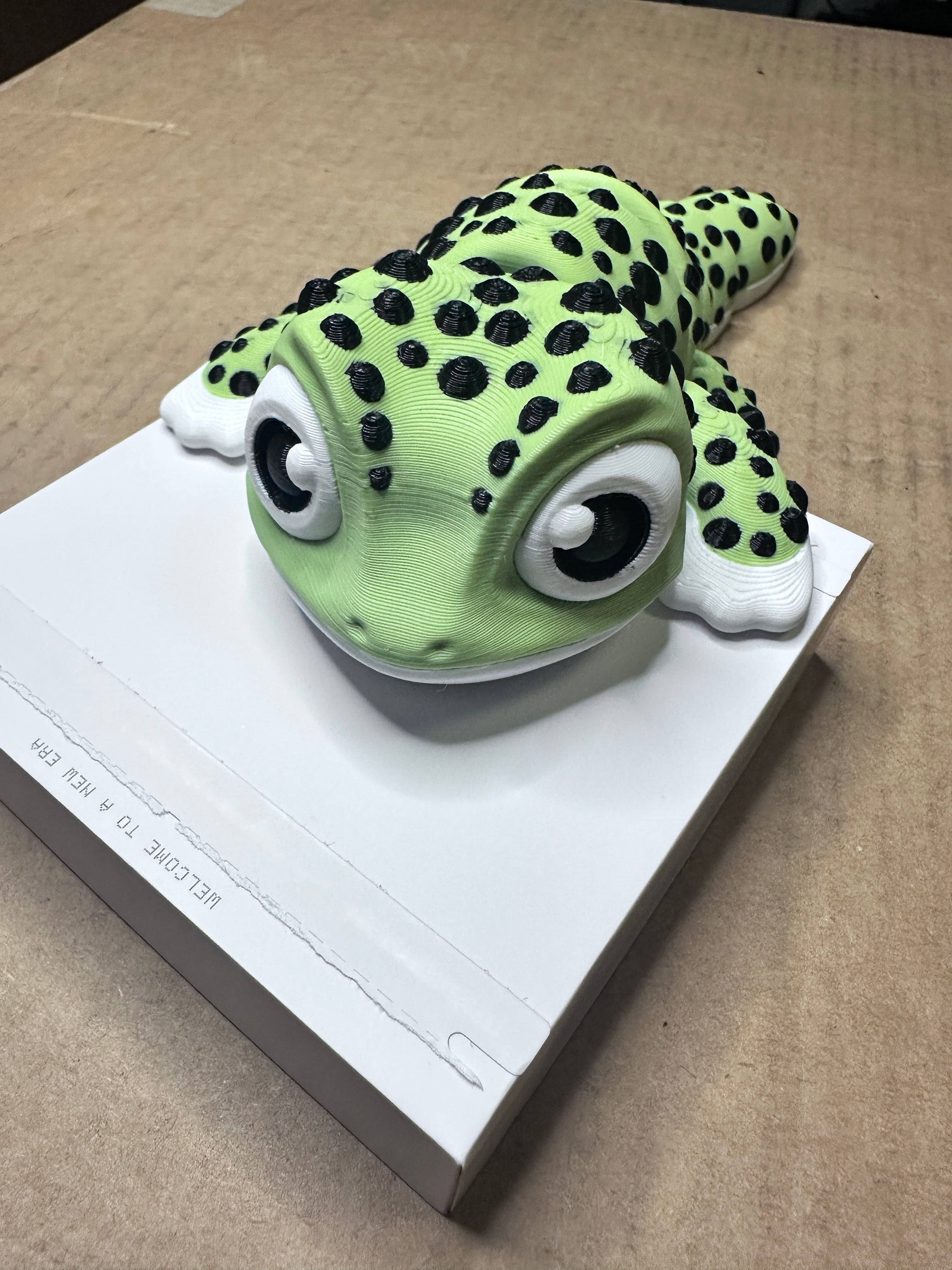 3D printed Gecko with a light green body, black spots, and large cartoon eyes, resting on a white rectangular box on a cardboard surface.