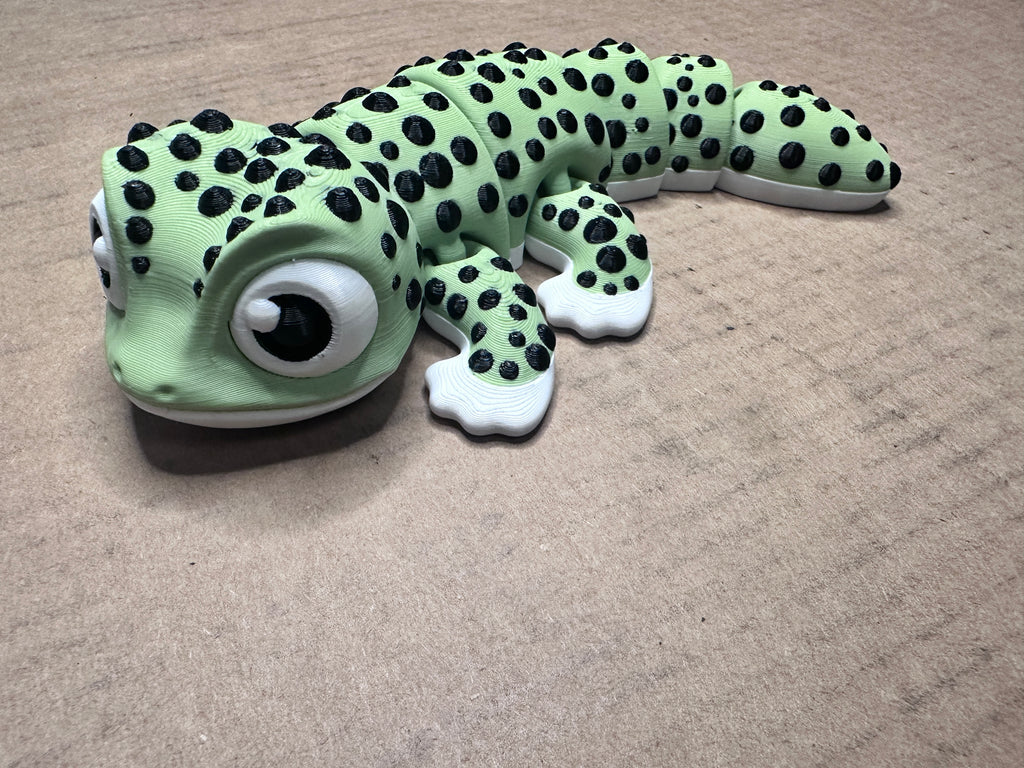 A green and black spotted 3D printed gecko with large cartoon eyes, curved slightly photographed on a beige background.