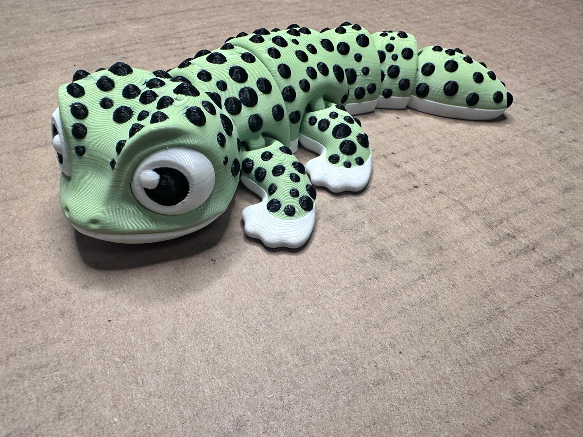 A green and black spotted 3D printed gecko with large cartoon eyes, curved slightly photographed on a beige background.