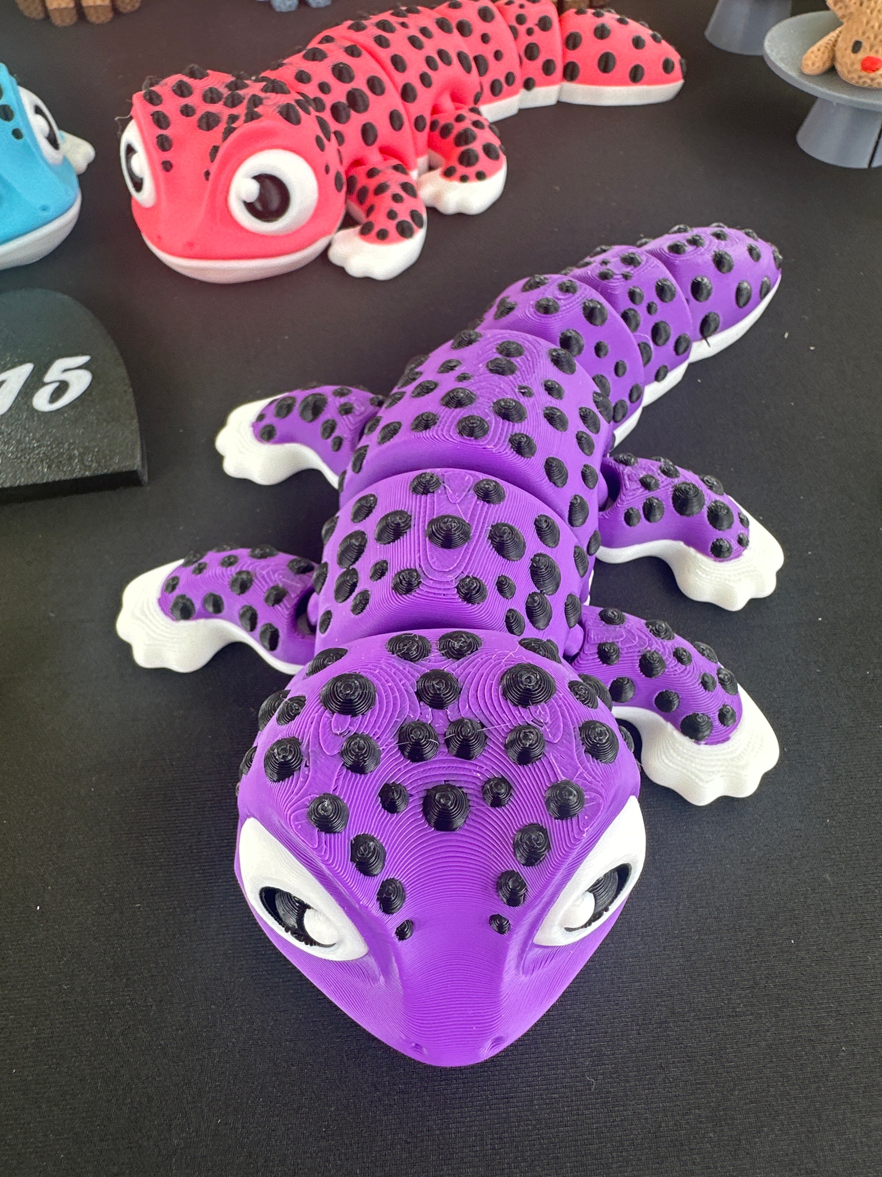 A purple 3D-printed articulated gecko toy with black spots and white toes, displayed on a black desk alongside other a pink and blue geckos in the background