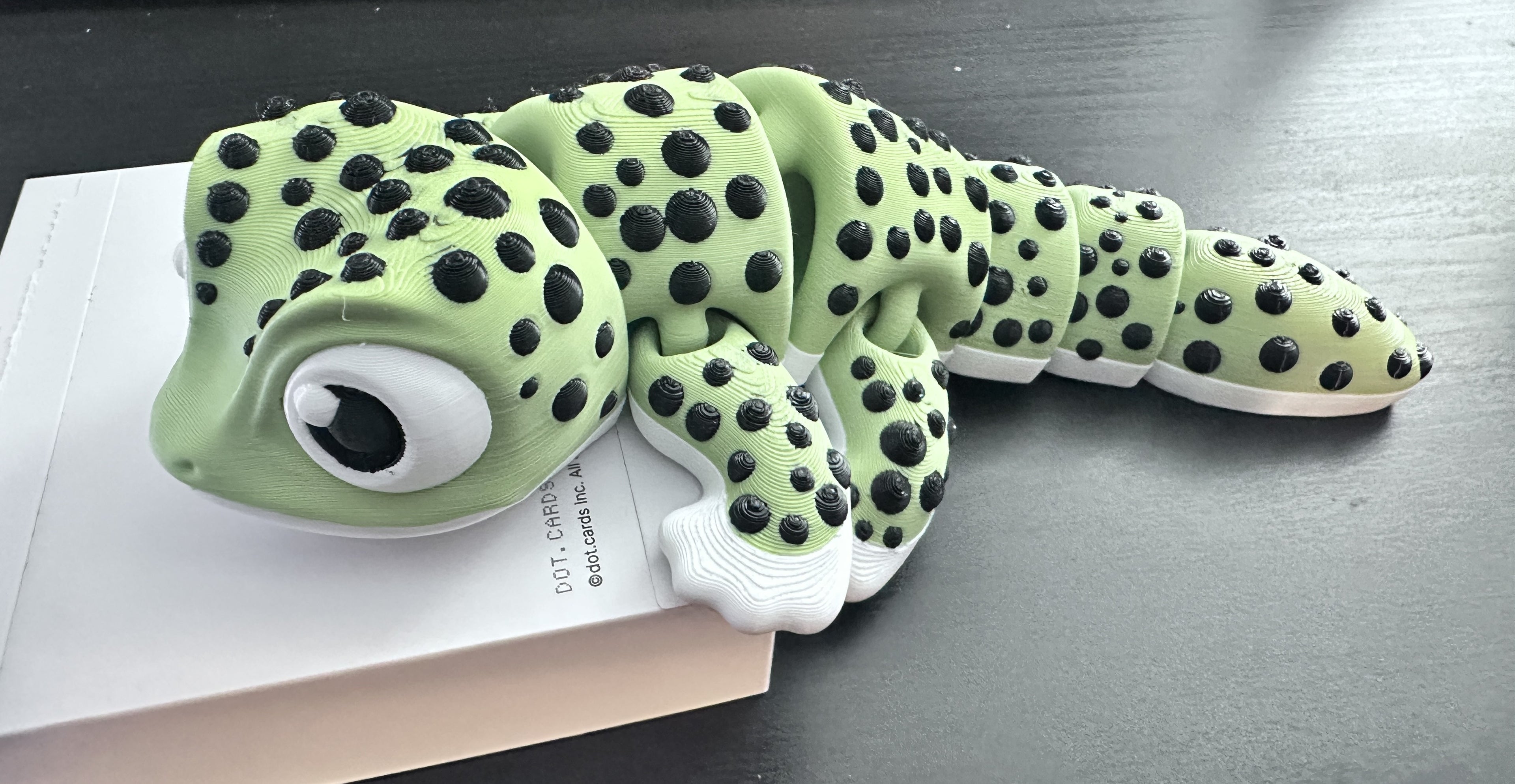 A green and black spotted gecko-3d printed articulated toy resting on top of a white box on a dark tabletop.