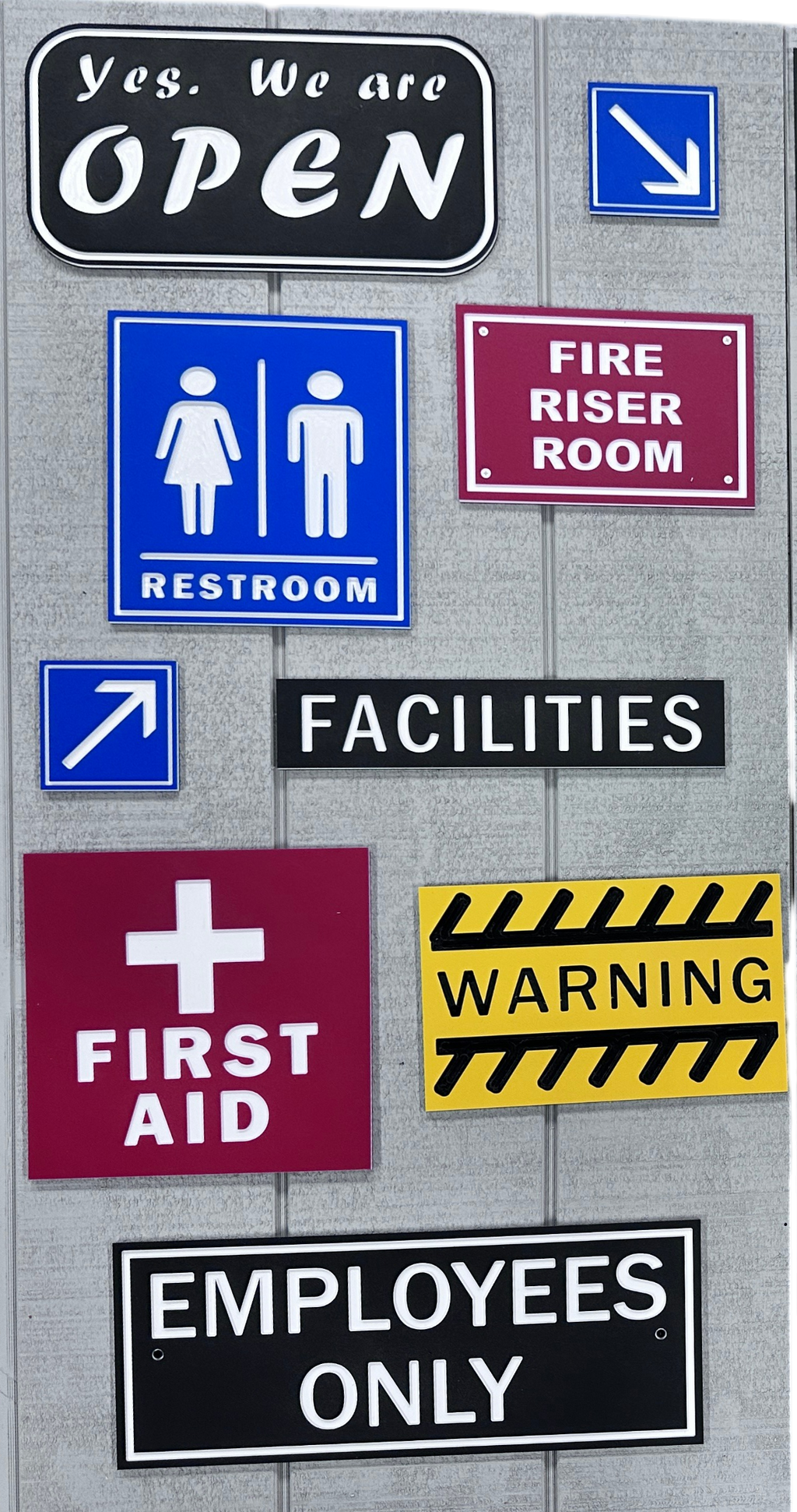 Collection of various signs indicating restroom, fire riser room, first aid, warning, and employees only on a gray wall.