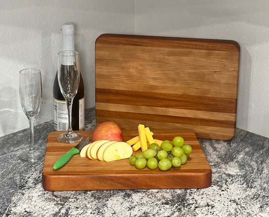 Mahogany block cutting board with cherry stripe displaying grapes, cheese, apple slices, bottle of wine and wineglasses