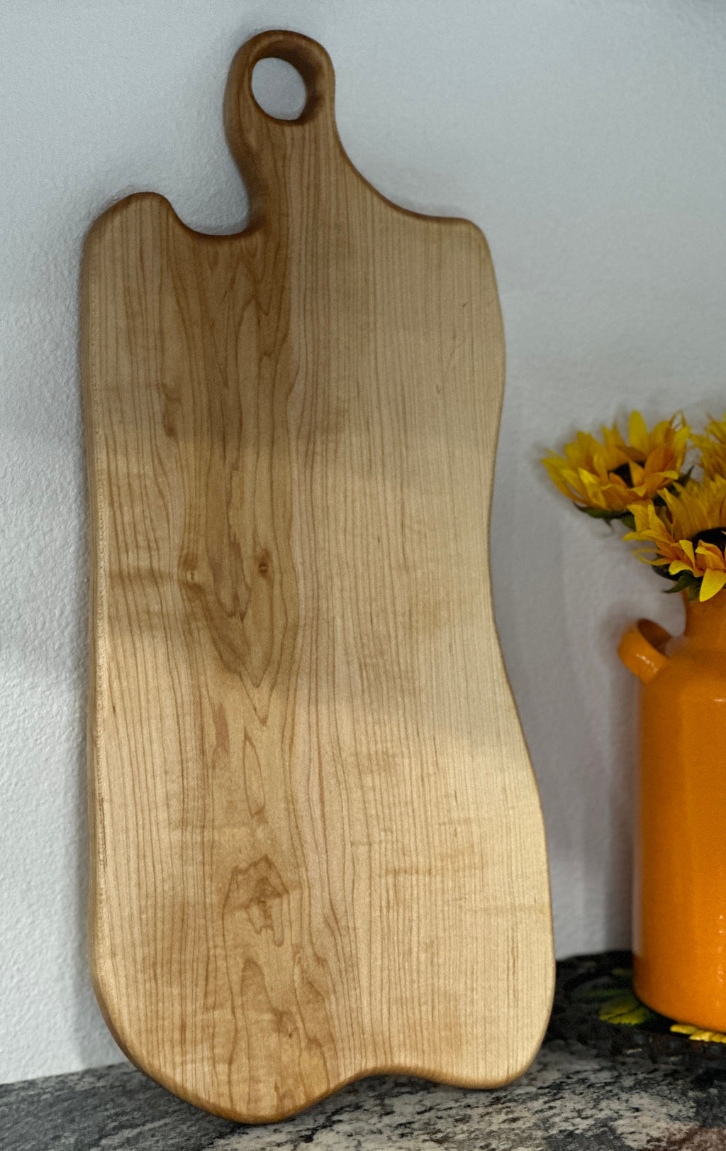 Maple light wood charcuterie board leaning against kitchen wall by an orange milk can vase with sunflowers