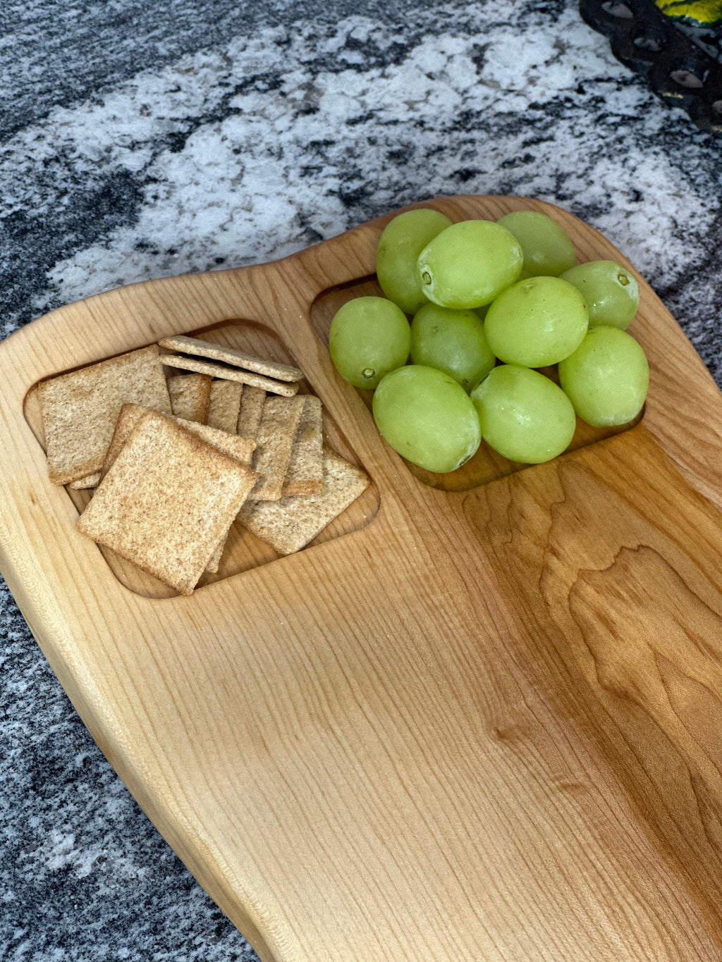 zoomed in view of Maple light wood charcuterie board with food sections filled with grapes and crackers on black and white marble countertop