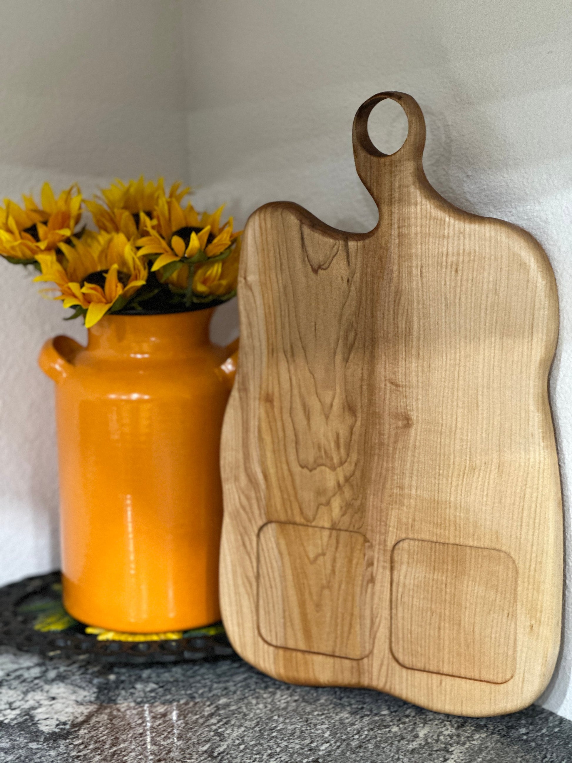 Maple light wood charcuterie board with food sections leaning against the wall next to an orange milk can vase with sunflowers