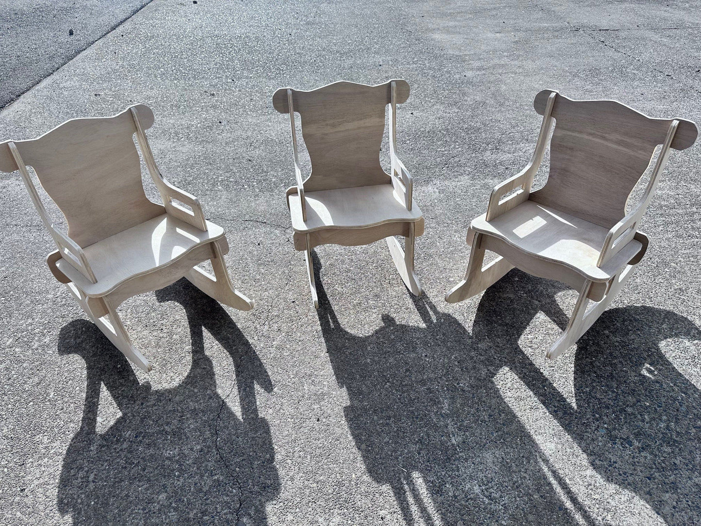 3 unfinished toddler rocking chairs outside with the sun casting shadows on the ground of each chair.