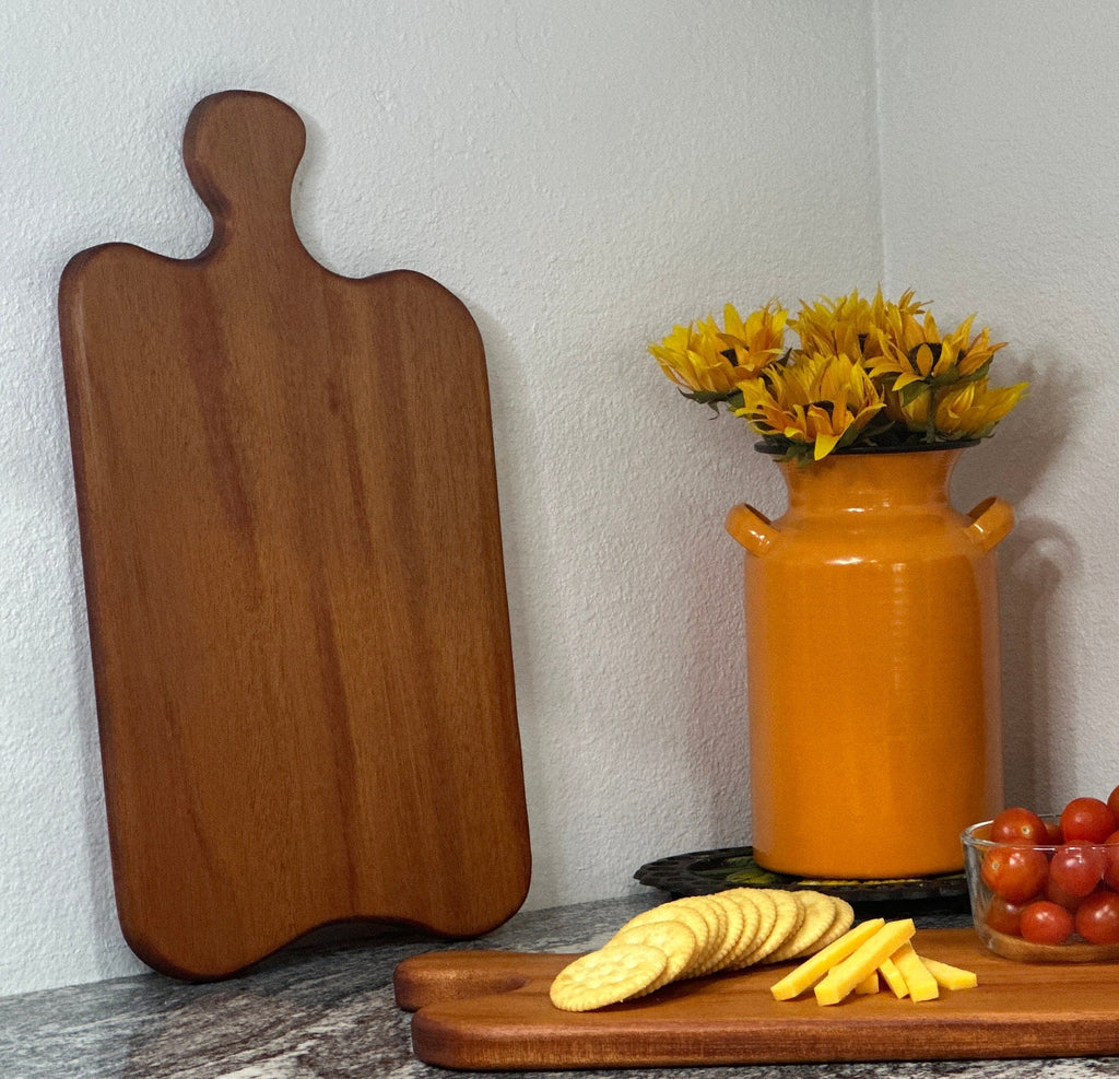 Mahogany red hardwood paddle shaped charcuterie board with cherry tomato, cheese and crackers displayed in front of an orange milk can vase with sunflowers