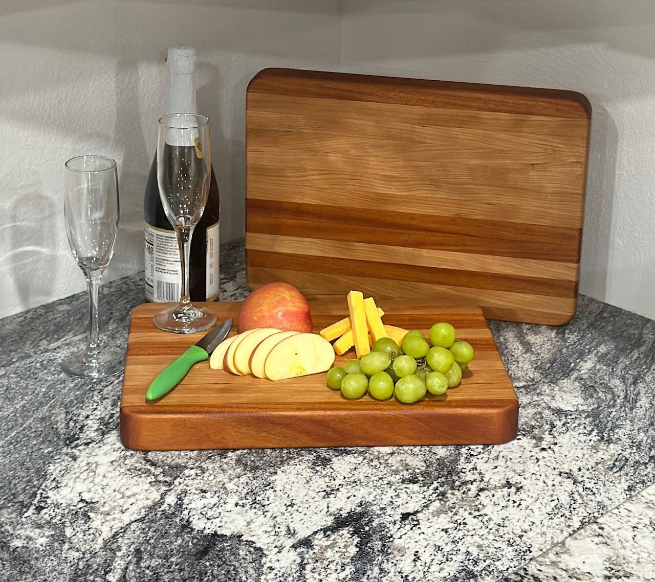 Mahogany block cutting board with cherry stripe displaying grapes, cheese, apple slices, bottle of wine and wineglasses