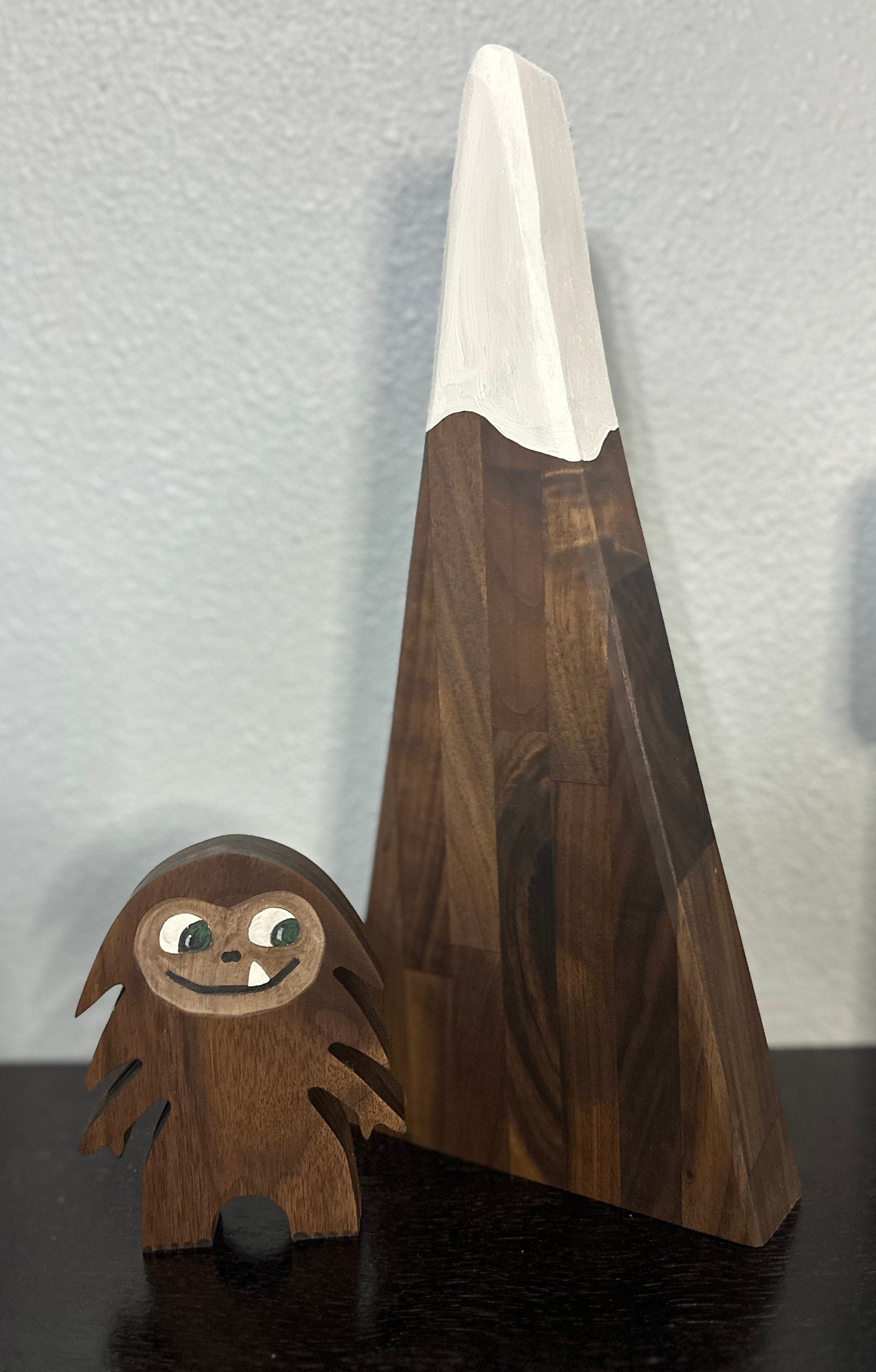 Hardwood small Bigfoot in walnut with smiling one-tooth mouth, background of dark triangle mountain with white painted snowcap