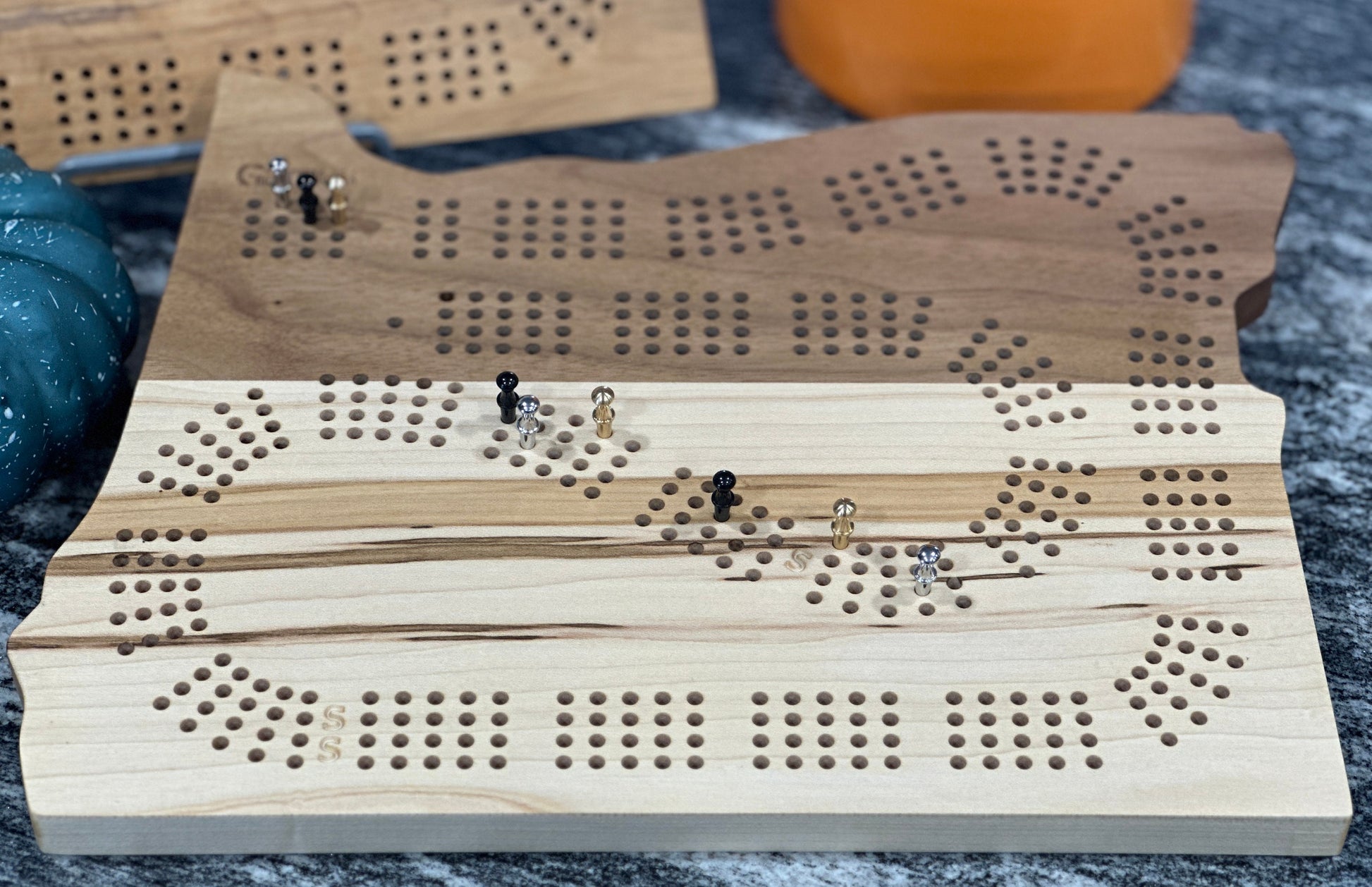 A zoomed in view of a 4player Oregon shaped cribbage boards made of maple and walnut with metal playing pins sitting on a grey/white marbled counter 