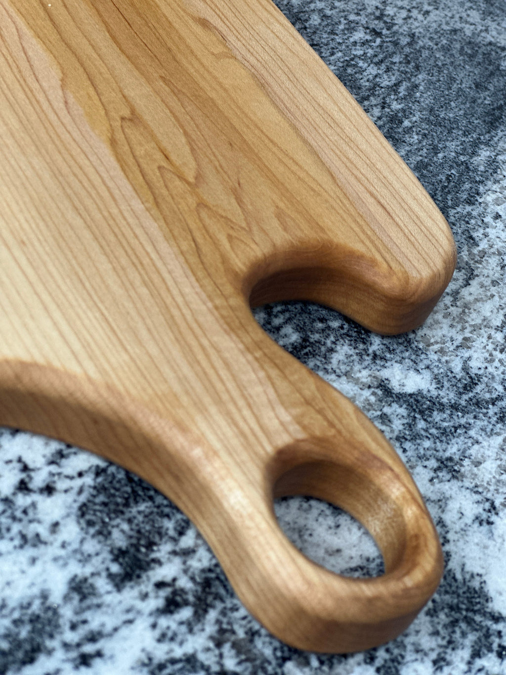 zoomed in view of handle with finger hole of Maple light wood charcuterie board on black and white countertop