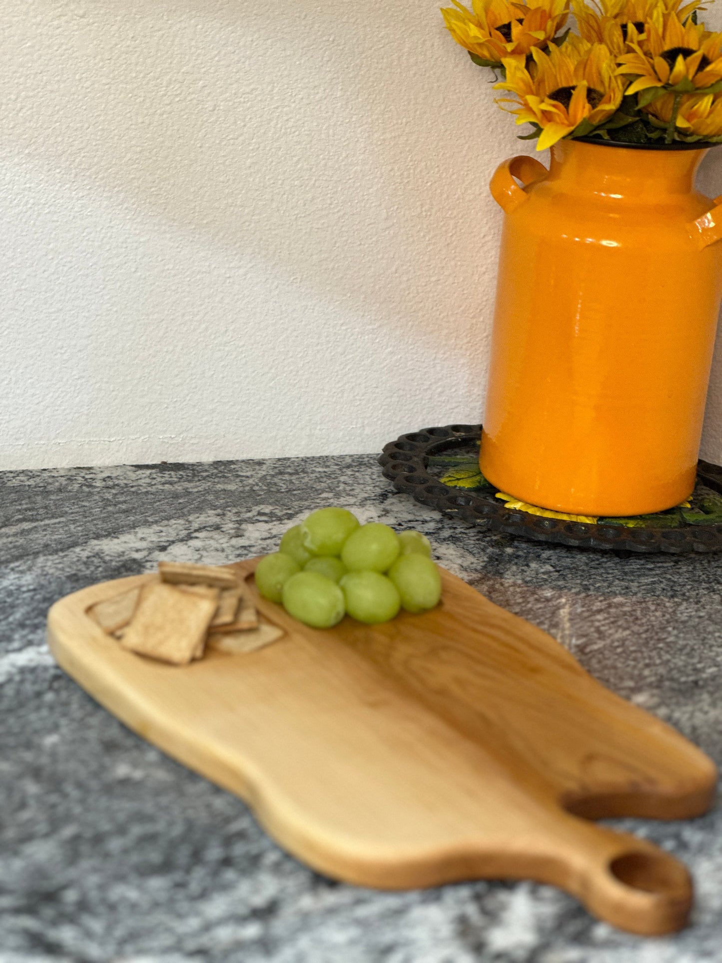 Maple light wood charcuterie board with food sections filled with grapes and crackers by an orange milk can vase with sunflowers