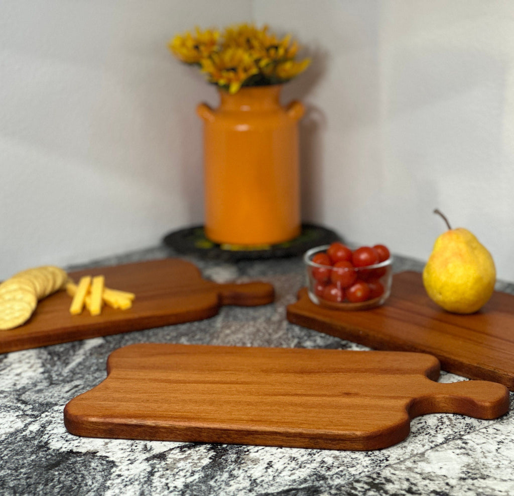 3 Mahogany red hardwood paddle shaped charcuterie boards with cherry tomato, a pear, cheese and crackers displayed in front of an orange milk can vase with sunflowers