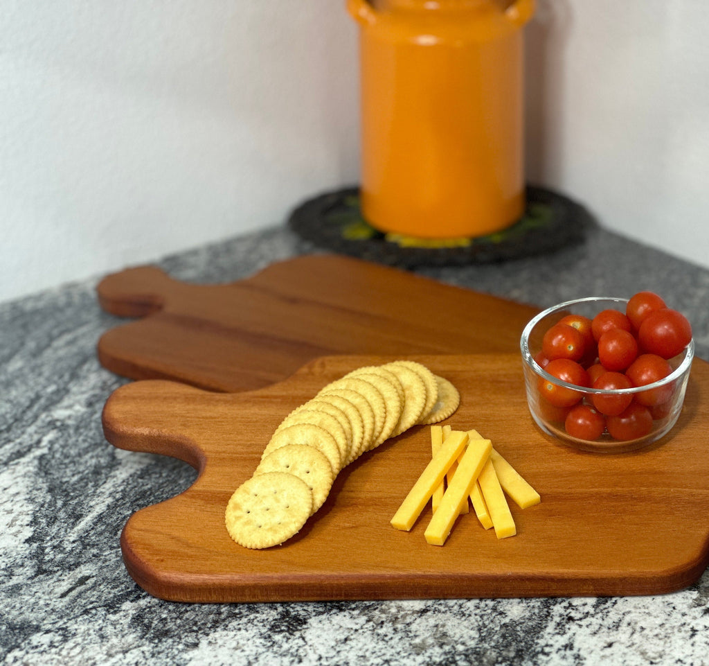 Mahogany red hardwood paddle shaped charcuterie board with cherry tomato, cheese and crackers displayed in front of an orange milk can vase