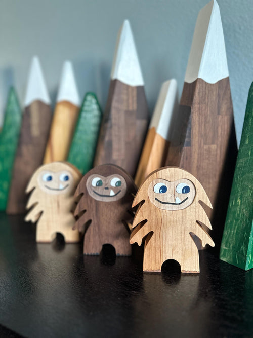 Hardwood Bigfoot in maple and walnut with smiling one-tooth mouth, background of light and dark triangle mountains with white painted snowcaps and green triangle trees
