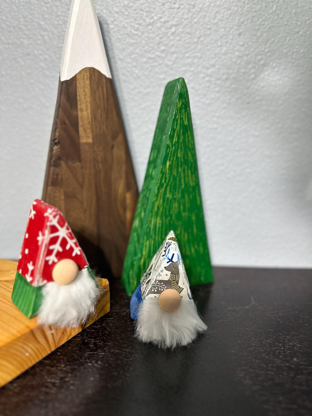 2 triangle gnomes with Christmas paper antiqued style hats sitting on a shelf with a wooden triangle walnut mountain and green tree in the background