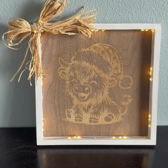 Video of lighted wooden picture frame with engraved design of a highland cow laughing and wearing a Santa hat, accented with a straw corner bow