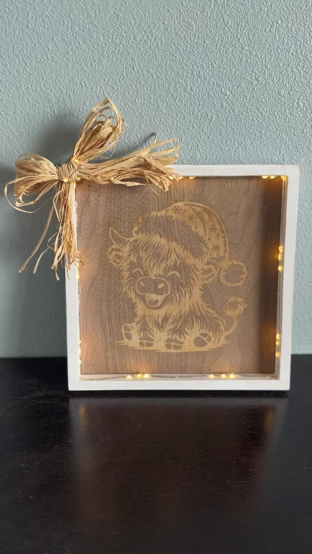 Video of lighted wooden picture frame with engraved design of a highland cow laughing and wearing a Santa hat, accented with a straw corner bow